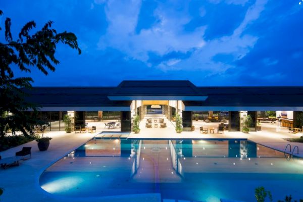 An illuminated outdoor pool area of a modern building at dusk, surrounded by seating and trees, under a partly cloudy sky.