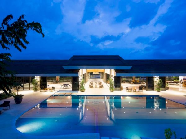 An illuminated outdoor pool area of a modern building at dusk, surrounded by seating and trees, under a partly cloudy sky.