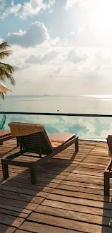 A serene poolside with lounge chairs, umbrellas, palm trees, and a view of the sea under a bright, partly cloudy sky.