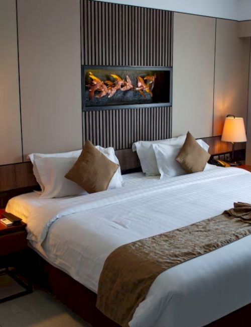 A cozy hotel room with a large bed, two nightstands with lamps, and a wall-mounted fireplace, decorated with neutral tones and warm lighting.