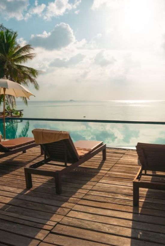 A serene poolside scene with lounge chairs, umbrellas, palm trees, and a tranquil water view under a partly cloudy sky.