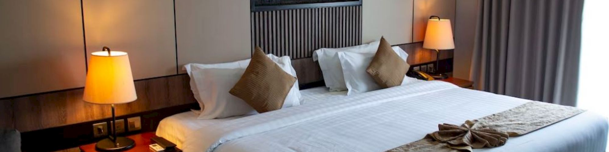A well-lit hotel room with a large bed, two side tables with lamps, decorative pillows, wall art, and curtains covering a window.