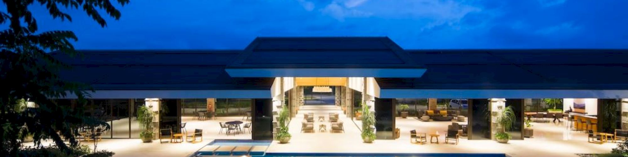 An outdoor pool area at dusk with a large, modern building in the background and lounge areas on both sides.