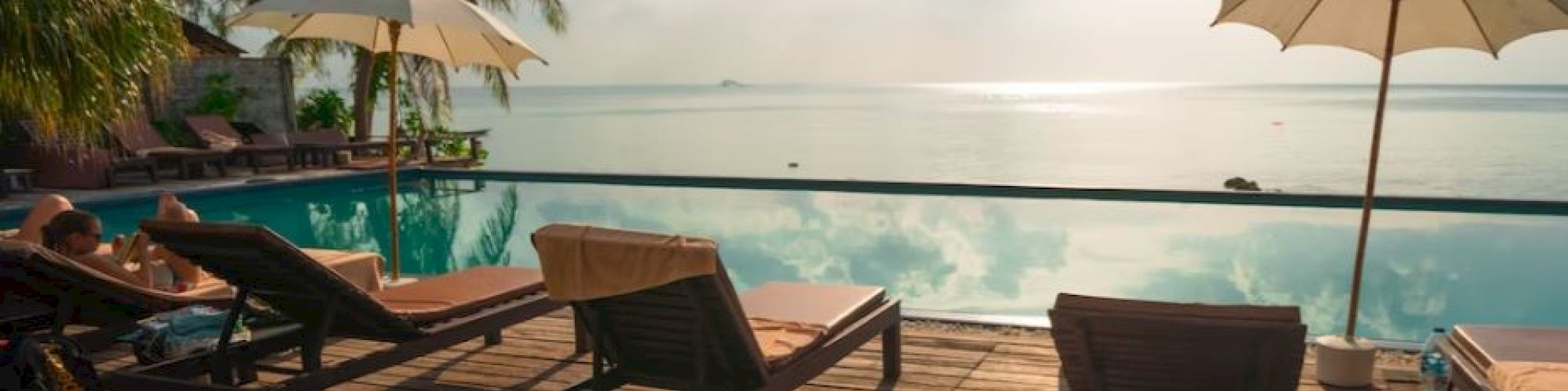 A tropical resort features sun loungers, sun umbrellas, and a wooden deck overlooking an infinity pool with a scenic ocean view at sunset.