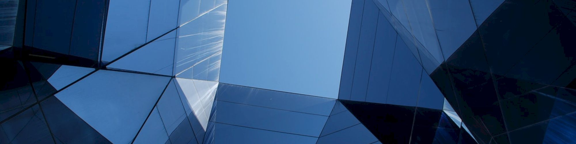 The image shows a geometric, architectural structure made of reflective glass panels, forming a skylight through which the clear blue sky is visible.