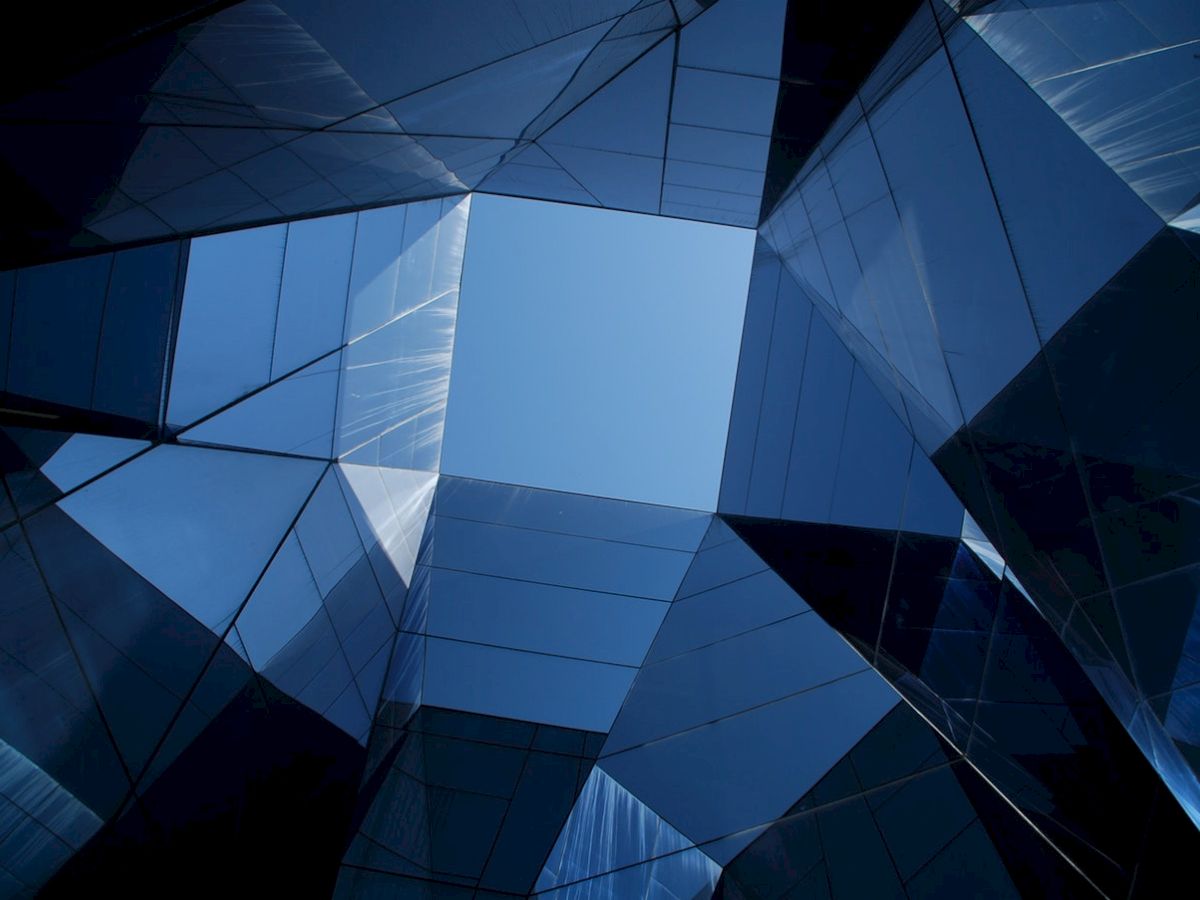 The image shows a view looking upward through a geometric arrangement of reflective, blue-tinted glass panels, revealing a patch of blue sky in the center.