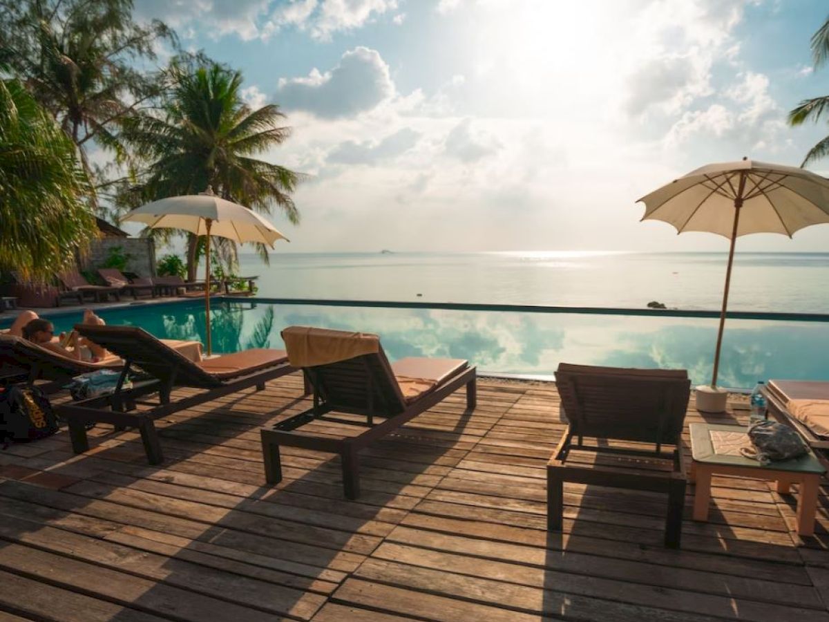 A serene poolside scene with lounge chairs, umbrellas, and a stunning ocean view, framed by palm trees, under a bright sky filled with sun and clouds.
