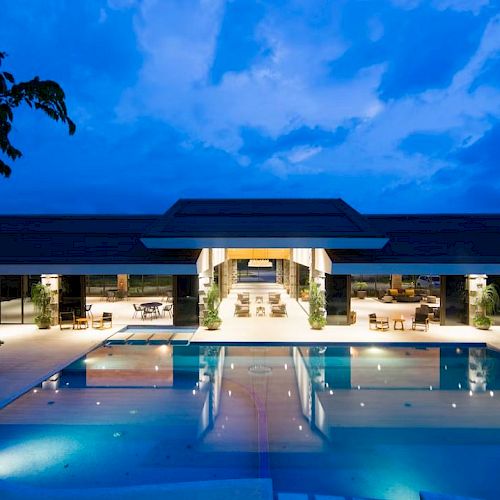 An illuminated, modern outdoor pool with a luxurious building surrounded by seating areas and a clear evening sky.