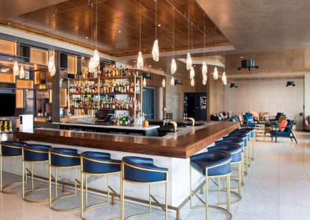 A modern bar with a wooden counter, blue barstools, hanging lights, and an assortment of bottles, located in a spacious lounge area.