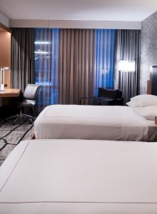 A hotel room with two neatly made beds, nightstands, a wall-mounted TV, a desk with a chair, and a large window with curtains.