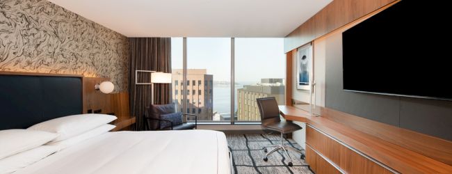 A modern hotel room with a large bed, a flat-screen TV, a desk, and floor-to-ceiling windows offering a city view.