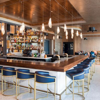 A modern bar with high stools, pendant lighting, and a fully stocked shelf of bottles behind the counter. A lounge area is visible in the background.