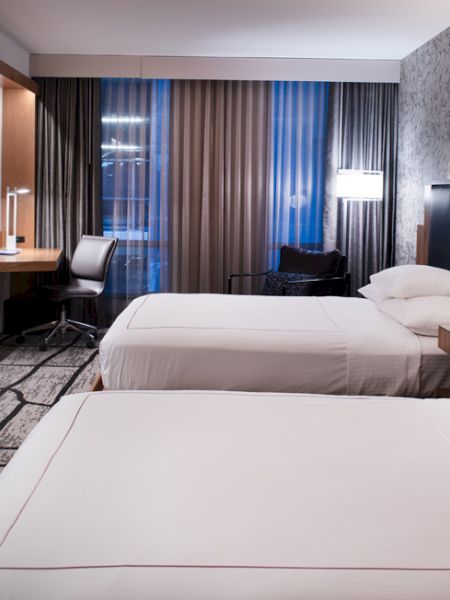 A modern hotel room with two beds, a wall-mounted TV, a desk with chair, and large windows with curtains and a city view outside.