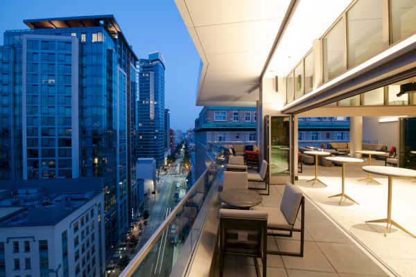 A modern rooftop dining area at dusk with city skyscrapers in the background, featuring tables and chairs, and an open indoor seating area.