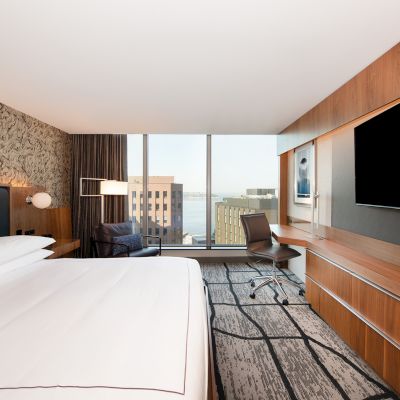 A modern hotel room with a large bed, wall-mounted TV, desk, chair, and a window offering a city and waterfront view.