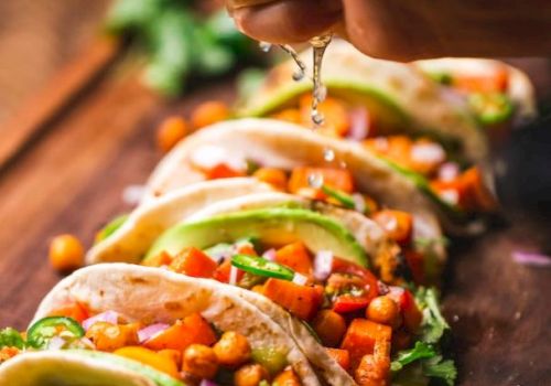 The image shows several tacos filled with chickpeas, vegetables, avocado slices, and garnished with fresh ingredients, being topped with lime juice.