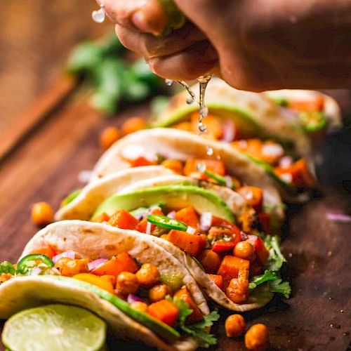 The image shows several tacos filled with chickpeas, vegetables, avocado slices, and garnished with fresh ingredients, being topped with lime juice.