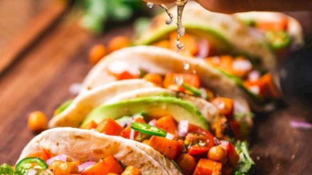 The image shows several tacos filled with chickpeas, vegetables, avocado slices, and garnished with fresh ingredients, being topped with lime juice.