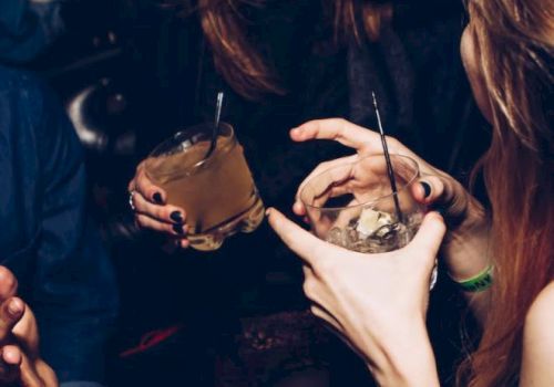 A group of people are socializing, holding drinks and laughing, likely in a dimly lit, festive setting or party environment.