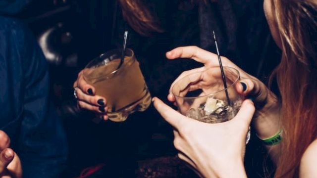 A group of people are socializing, holding drinks and laughing, likely in a dimly lit, festive setting or party environment.