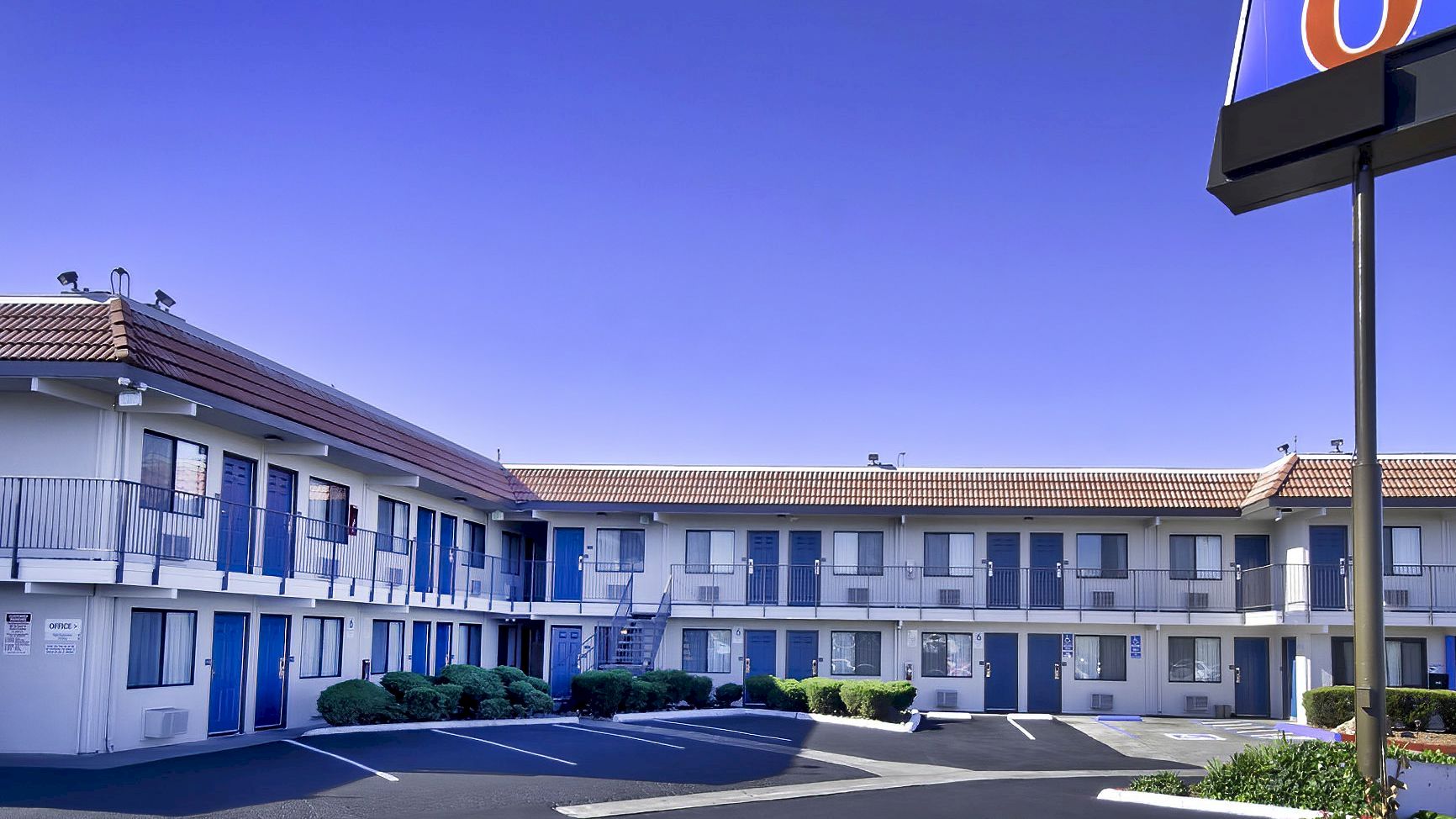 A two-story motel with blue doors and a prominent sign is shown. The parking lot is mostly empty, and the sky is clear.