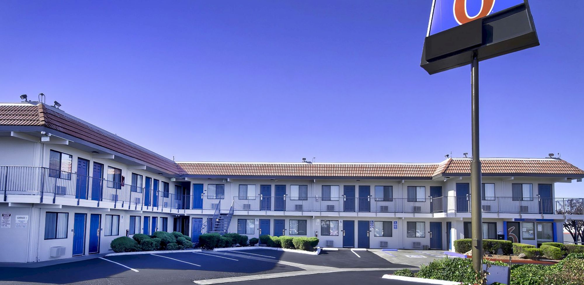 A two-story motel with blue doors and a prominent sign is shown. The parking lot is mostly empty, and the sky is clear.