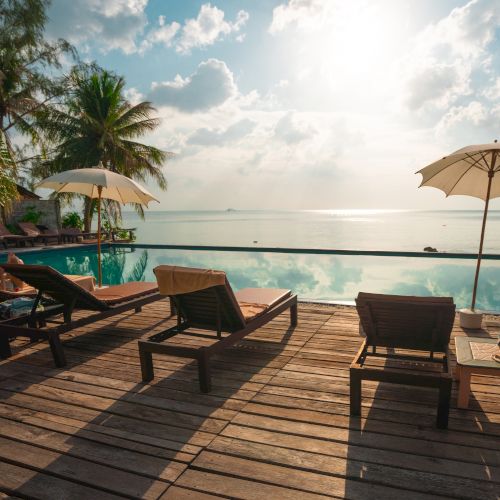 A serene poolside setting with wooden lounge chairs, umbrellas, palm trees, and a view of the ocean at sunset.
