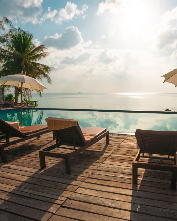 A serene poolside setting with wooden lounge chairs, umbrellas, palm trees, and a view of the ocean at sunset.