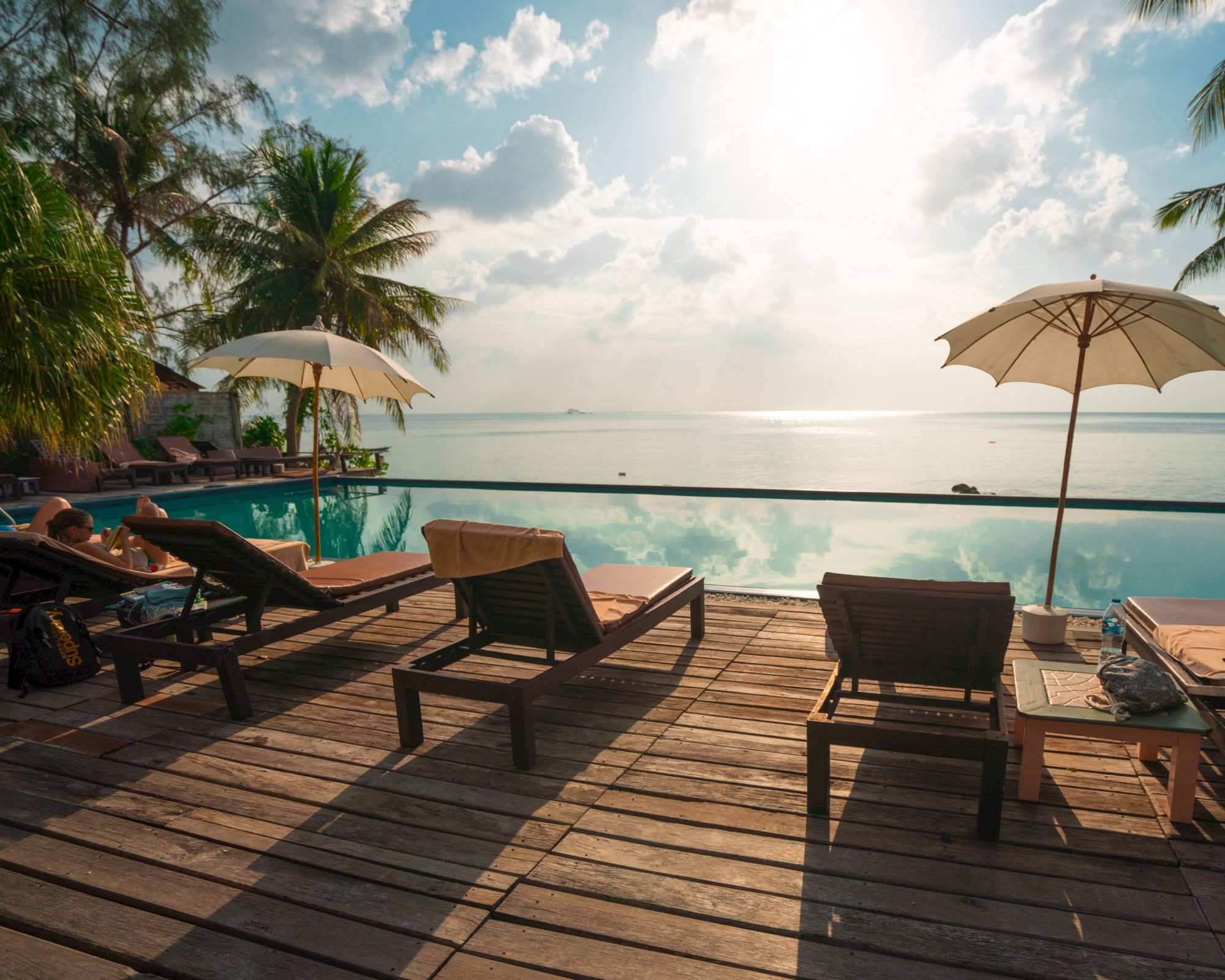 Sun loungers and umbrellas by a pool, overlooking a calm sea with a serene sky, surrounded by lush palm trees.