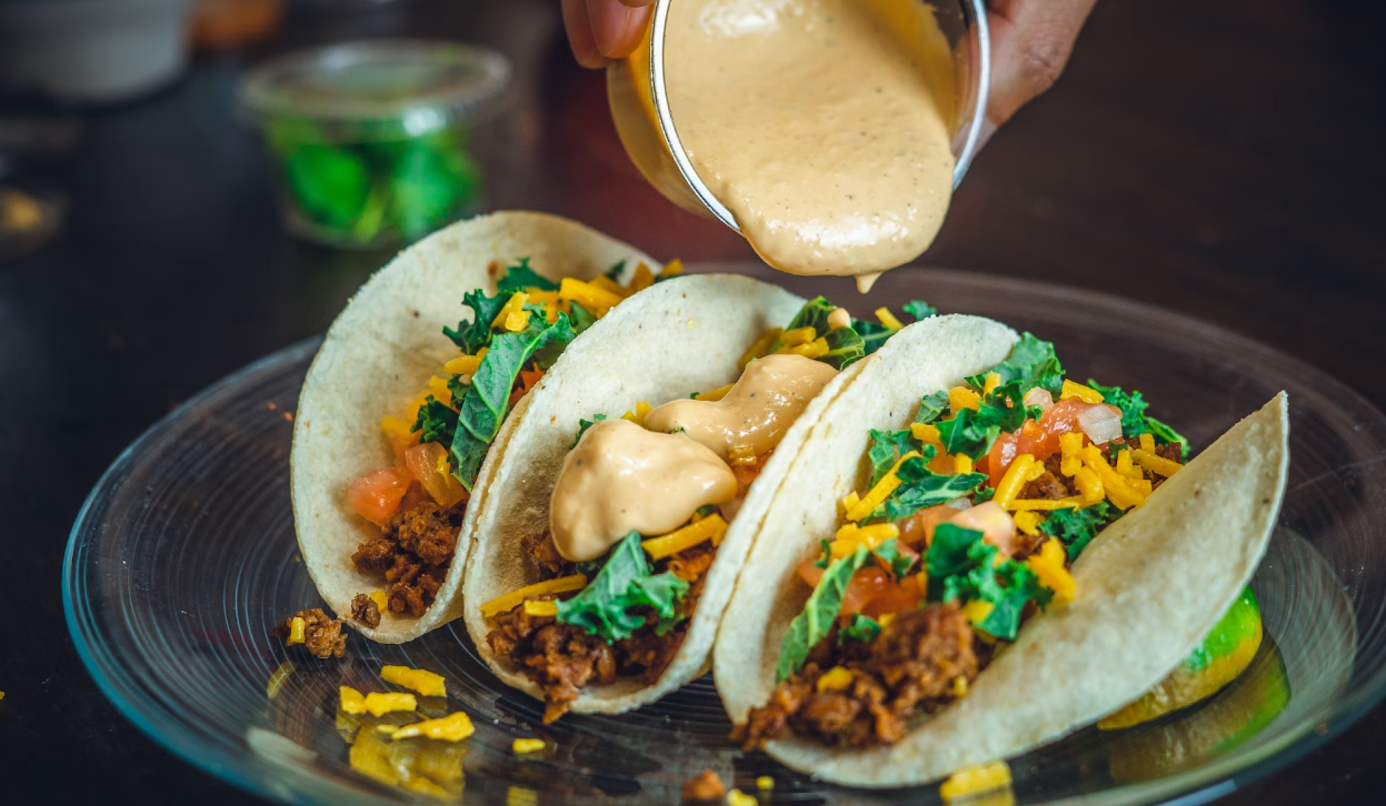 Three tacos filled with what appears to be ground meat, shredded cheese, greens, and other toppings, with sauce being poured over them.