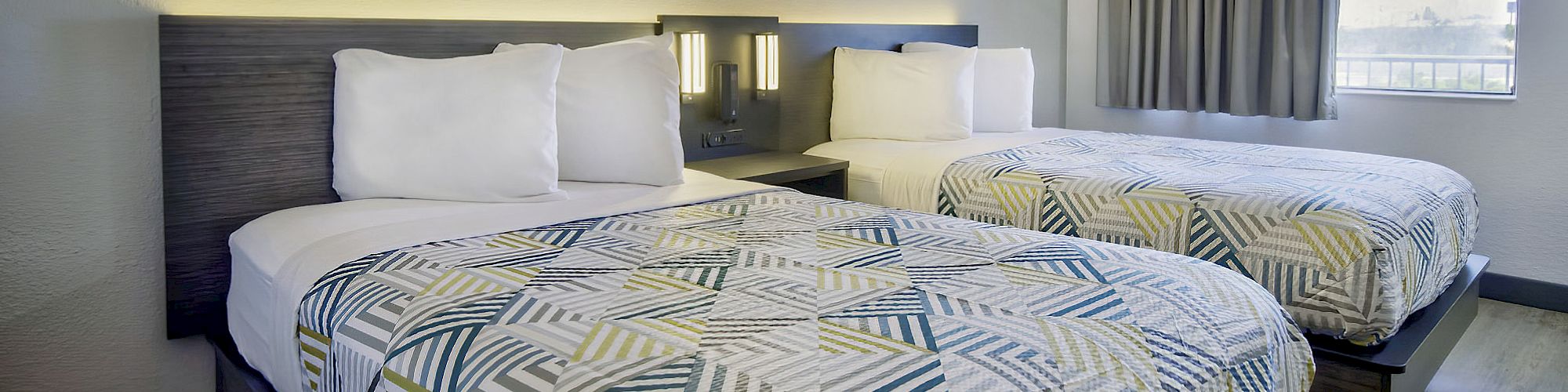 The image shows a modern hotel room with two double beds, patterned bedspreads, a nightstand between them, and a large window with curtains.