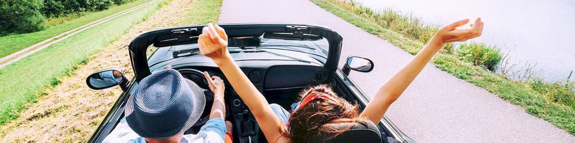 Two people in a convertible car driving along a river, one person's hands are up in the air, with scenic mountains and greenery in the background.