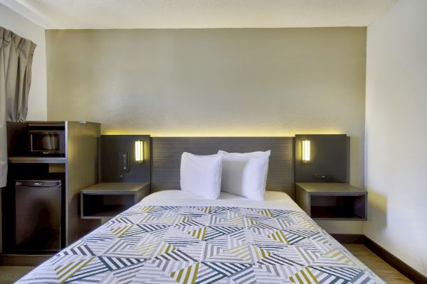 A neatly made bed with white pillows and a geometric-patterned cover in a modern room; side tables with lights, and a mini-fridge and microwave adjacent.