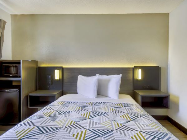 A neatly made bed with white pillows and a geometric-patterned cover in a modern room; side tables with lights, and a mini-fridge and microwave adjacent.