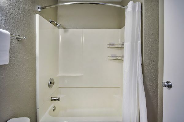 This image shows a clean, white bathtub and shower combo with a white curtain, modern fixtures, a towel on a rack, and small shelves for toiletries.