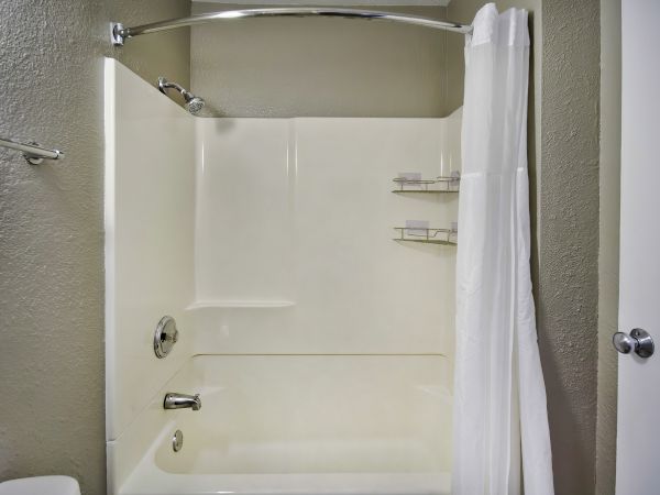 This image shows a clean, white bathtub and shower combo with a white curtain, modern fixtures, a towel on a rack, and small shelves for toiletries.