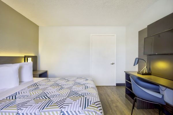 This image shows a neatly arranged bedroom with a bed, nightstand, desk, chair, lamp, and TV mounted on the wall, featuring modern furniture.