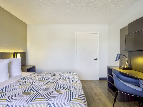 This image shows a neatly arranged bedroom with a bed, nightstand, desk, chair, lamp, and TV mounted on the wall, featuring modern furniture.