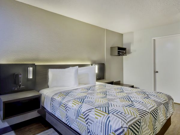 A modern hotel room with a neatly made bed, geometric patterned comforter, nightstands, wall-mounted lights, and a small shelf near the door.