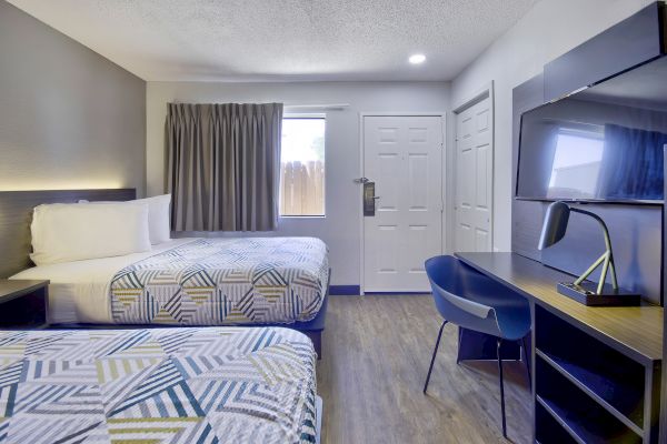 The image shows a hotel room with two neatly made beds, a desk with a chair, a TV, and a window with closed curtains.