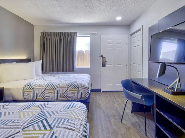 The image shows a hotel room with two neatly made beds, a desk with a chair, a TV, and a window with closed curtains.