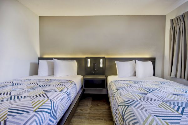 The image depicts a hotel room with two twin beds, featuring patterned bedspreads and white pillows, with a nightstand and wall lighting between the beds.