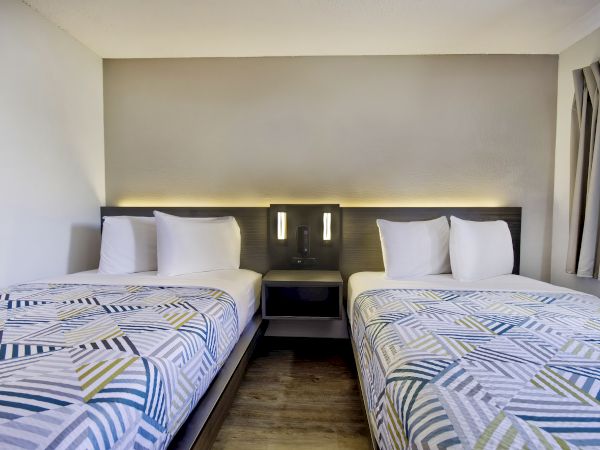 The image depicts a hotel room with two twin beds, featuring patterned bedspreads and white pillows, with a nightstand and wall lighting between the beds.