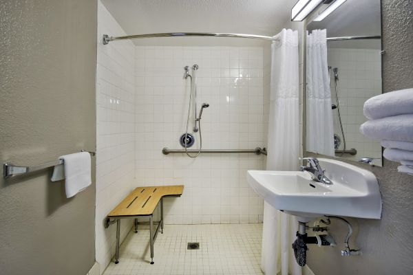 The image shows an accessible bathroom with a shower bench, grab bars, sink, and towels. The bathroom has white tiles and a shower curtain.