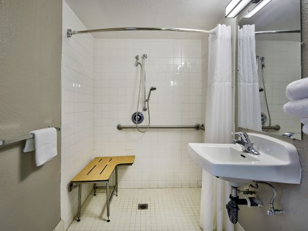 The image shows an accessible bathroom with a shower bench, grab bars, sink, and towels. The bathroom has white tiles and a shower curtain.