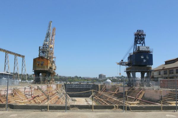 The image shows an industrial shipyard with cranes, fenced off sections, and a clear sky in the background.