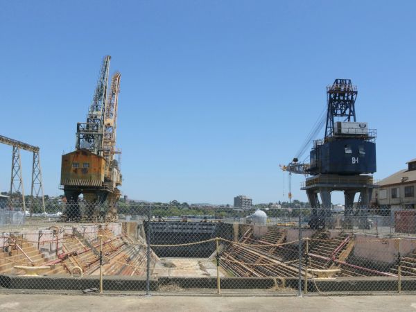 The image shows an industrial shipyard with cranes, fenced off sections, and a clear sky in the background.