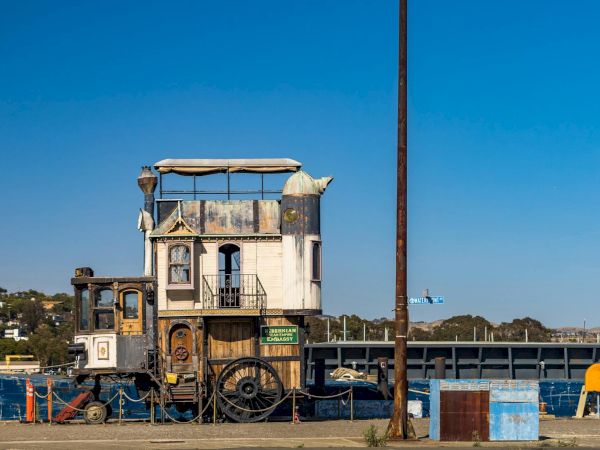 A whimsical, old-fashioned houseboat with an eclectic design is docked at a marina, featuring a mix of traditional and rustic elements.