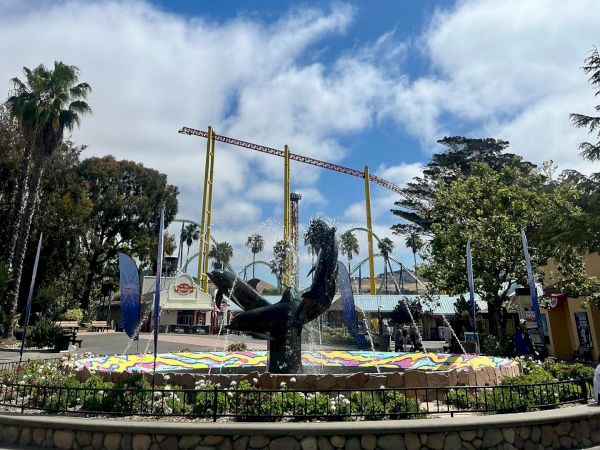The image shows a public square with a statue in the center, surrounded by colorful decorations, palm trees, and some buildings in the background.