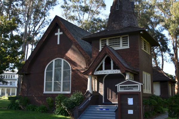 Image of a quaint brown church with a steep-roofed steeple, gothic windows, and a signboard in front, surrounded by trees and green grass.